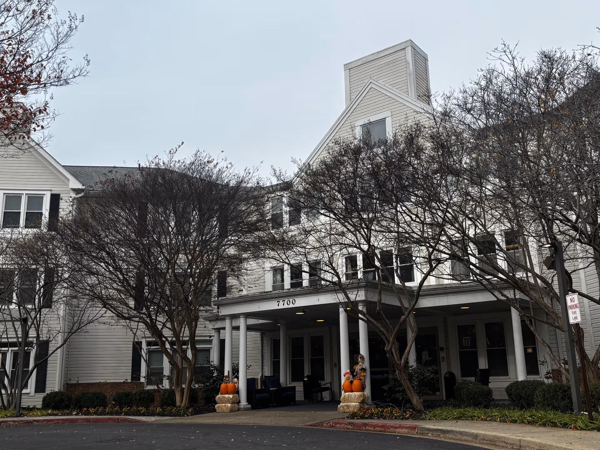 Exterior view of Morningstar Assisted Living facility showing a white multi-story building with black shutters, a covered entrance with columns, and autumn decorations including pumpkins and hay bales near the entrance. Leafless trees and landscaping surround the building under an overcast sky.