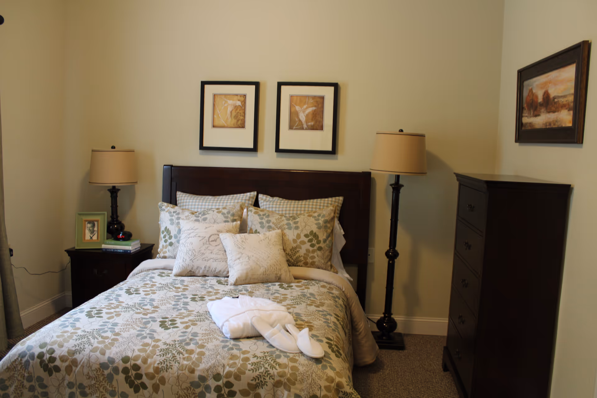 A neatly made bed with floral patterned bedding and multiple pillows in a bedroom. There are two framed pictures hanging above the dark wooden headboard. On the left side of the bed is a nightstand with a lamp, a framed photo, and some books. On the right side of the bed is a tall dark wooden dresser and a floor lamp. The walls are painted light beige.