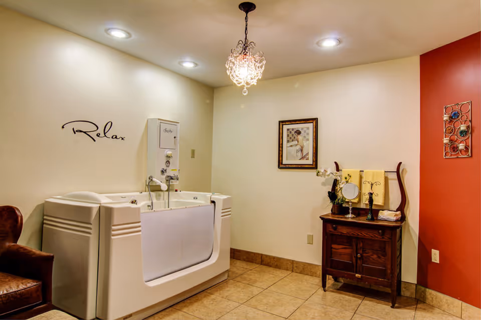 A serene room featuring a walk-in bathtub with the word 'Relax' on the wall above it. The room has beige walls with one red accent wall, a wooden cabinet with a mirror and towels, a framed picture, a decorative wall piece, a leather chair, and a small chandelier hanging from the ceiling.