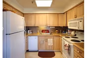 A compact kitchen with light wood cabinets, a white refrigerator, dishwasher, microwave, and electric stove. The countertop has various small kitchen items, and two beige towels hang on the dishwasher handle. A small brown rug is placed on the floor in front of the sink area.