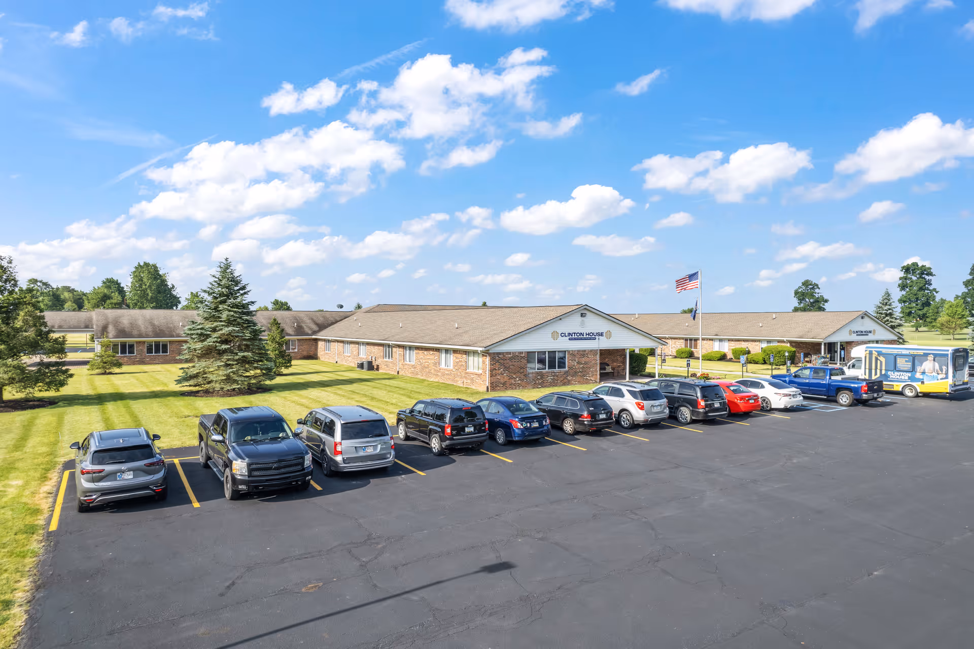 Exterior view of Clinton House Rehabilitation and Healthcare Center, a single-story brick building with a large parking lot in front. Several cars are parked, and there is an American flag flying on a flagpole near the entrance. The sky is blue with scattered clouds, and there are green trees and grass surrounding the building.