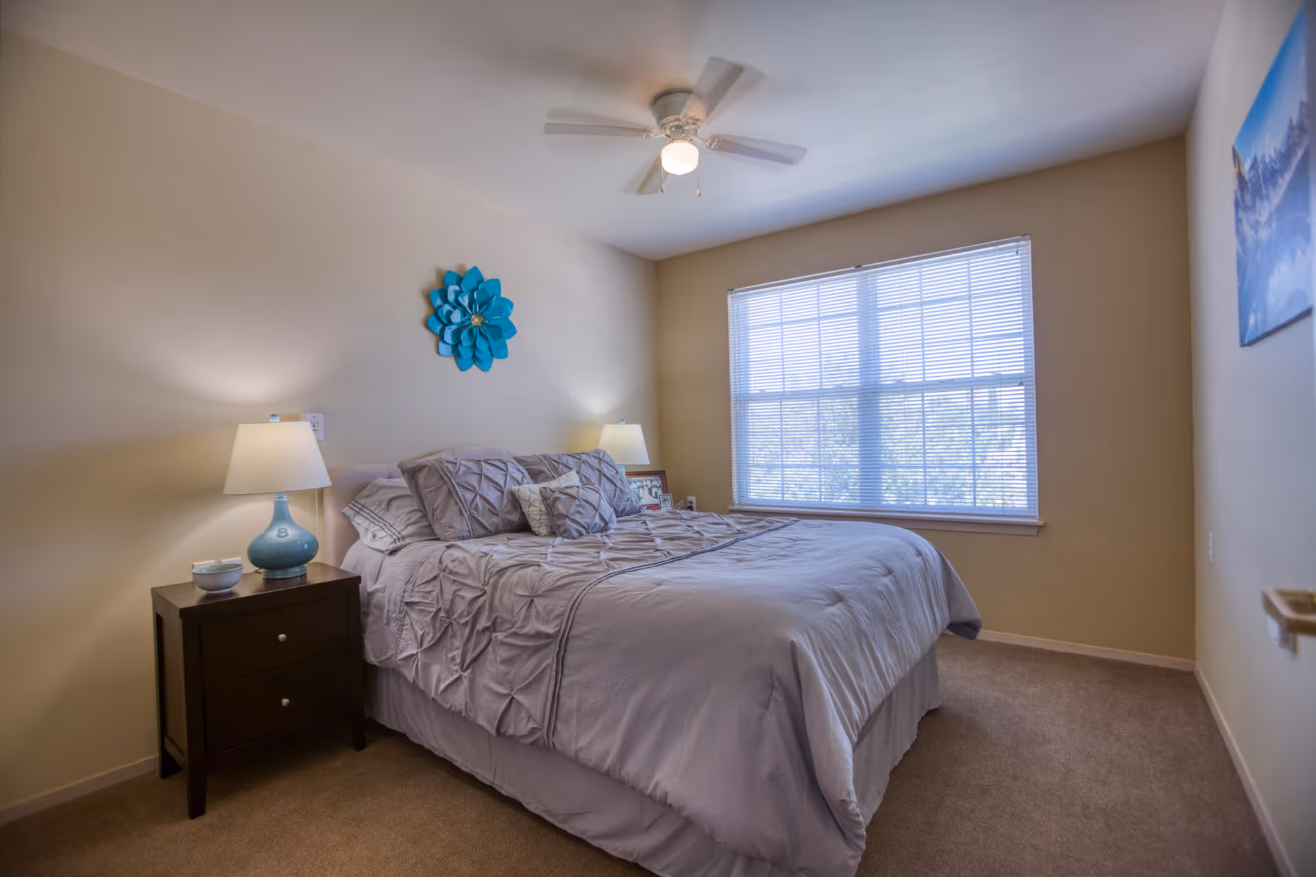 Sunlit bedroom with a made bed, two nightstands and lamps, a large window, ceiling fan, and turquoise wall decor.