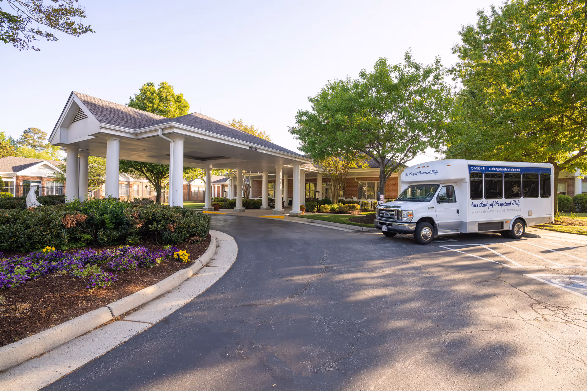 Covered entrance and porte-cochère of a senior living facility with a parked shuttle van and landscaped flowerbeds.