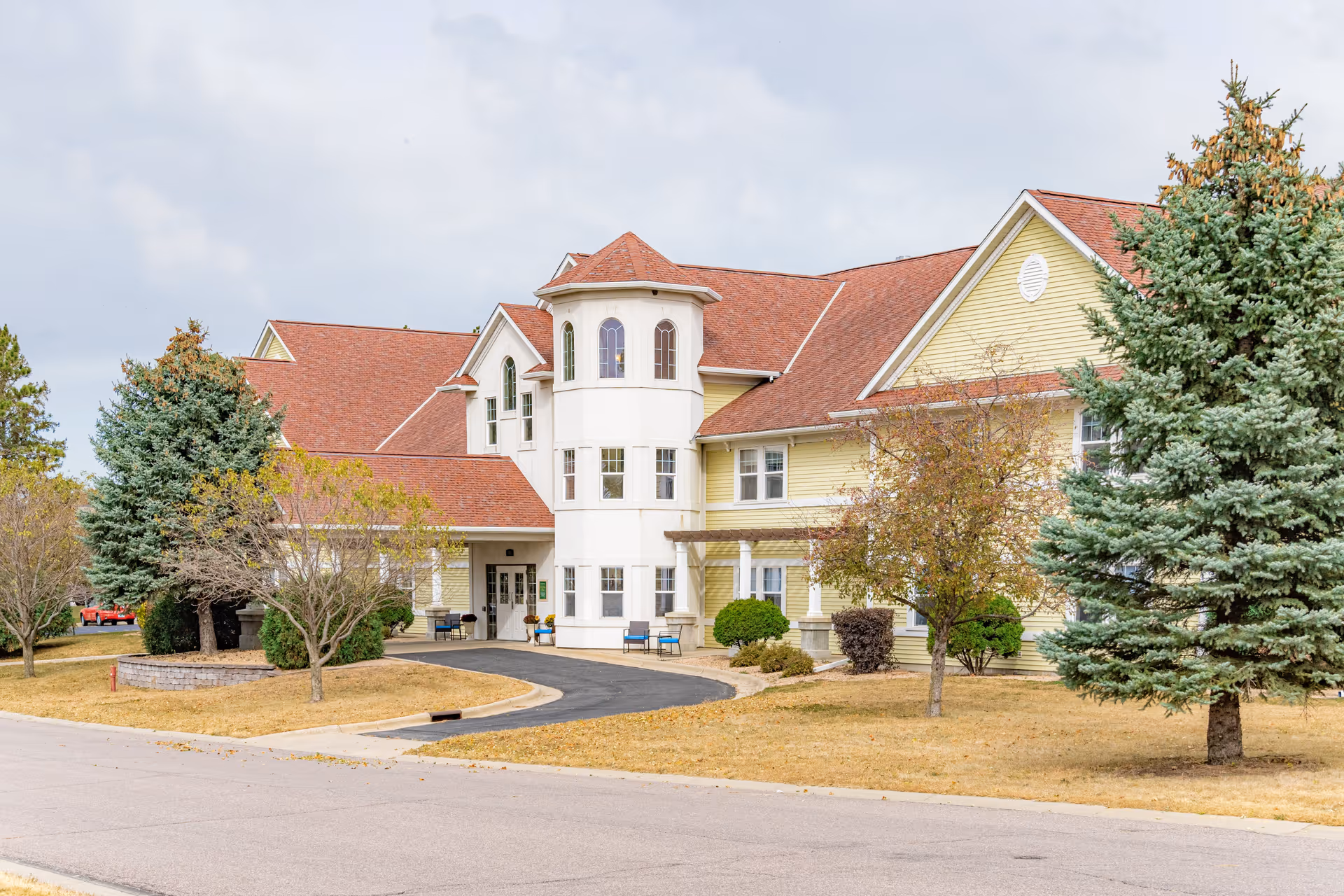 Exterior view of a large senior living facility building with yellow siding and a red roof, surrounded by trees and a driveway leading to the entrance.