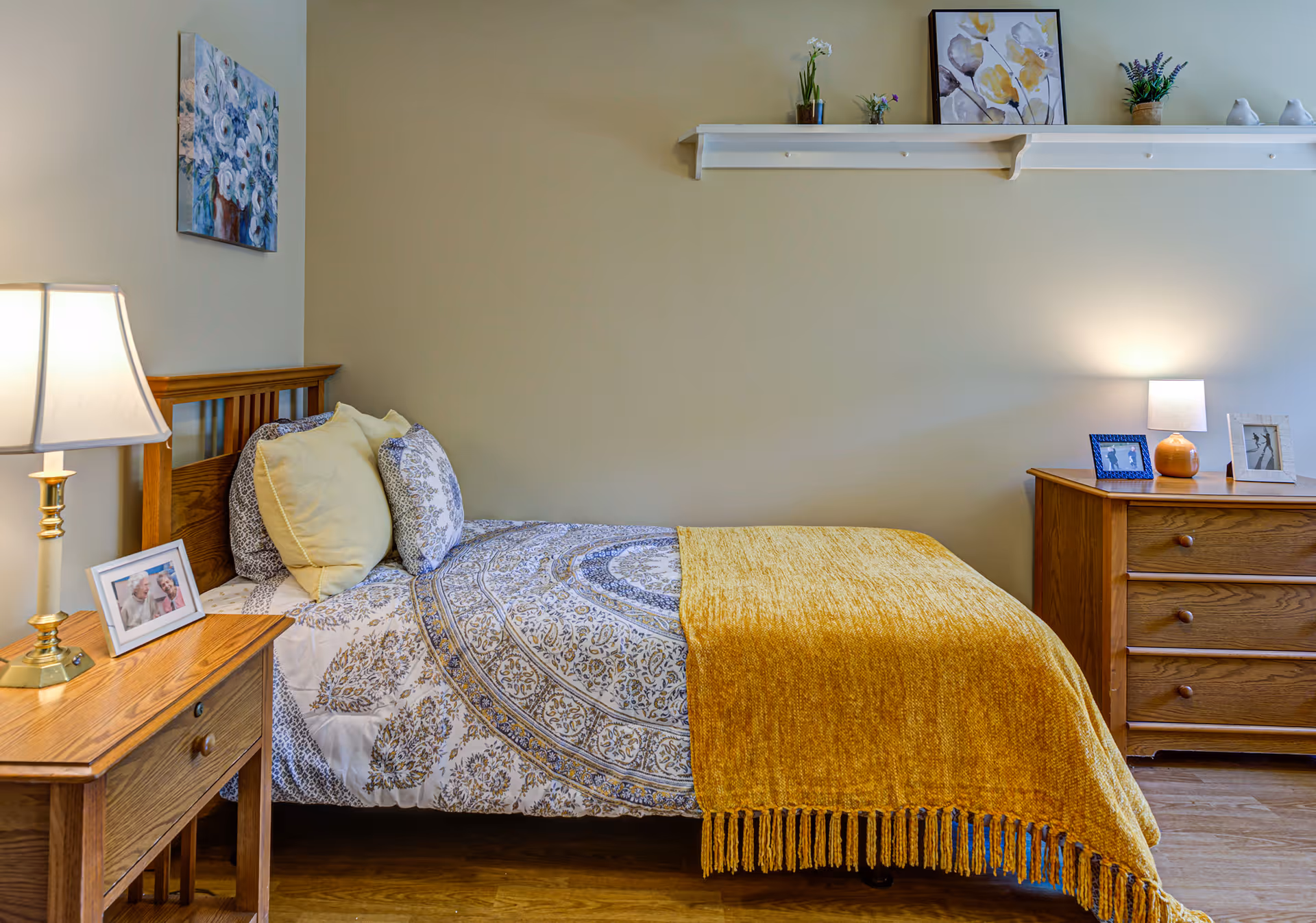 A cozy bedroom with a wooden bed frame, patterned bedding, and a mustard yellow throw blanket. There are two pillows on the bed, a wooden nightstand with a lamp and a framed photo, and a wooden dresser with a lamp and framed photos. A white shelf above the bed holds small plants and decorative items, and a floral painting hangs on the wall.