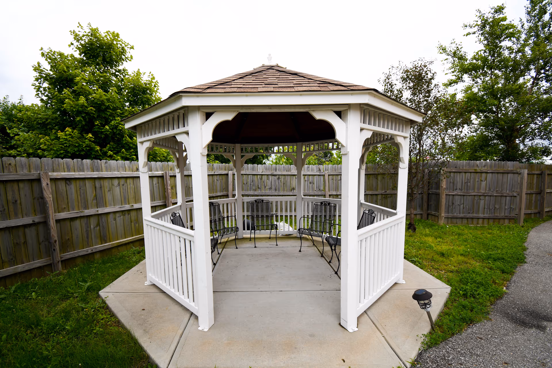 A white wooden gazebo with a shingled roof situated on a concrete pad in a fenced backyard area. Inside the gazebo are several black metal chairs arranged around the perimeter. Green grass and trees surround the gazebo, and a paved pathway is visible to the right.