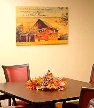 A dining area with a wooden table surrounded by red cushioned chairs. On the table is a decorative centerpiece made of autumn leaves and berries. On the wall above the table hangs a wooden artwork depicting a red barn with the text: 'God looked down on his planned paradise and said I need a caretaker SO GOD MADE A Farmer.'