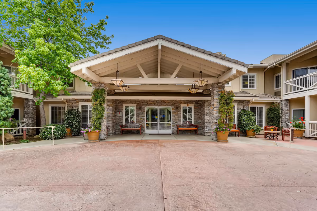 Entrance of Meadowbrook of Agoura Hills senior living facility featuring a covered driveway with stone pillars, hanging lantern-style lights, benches, potted plants, and a clear blue sky.