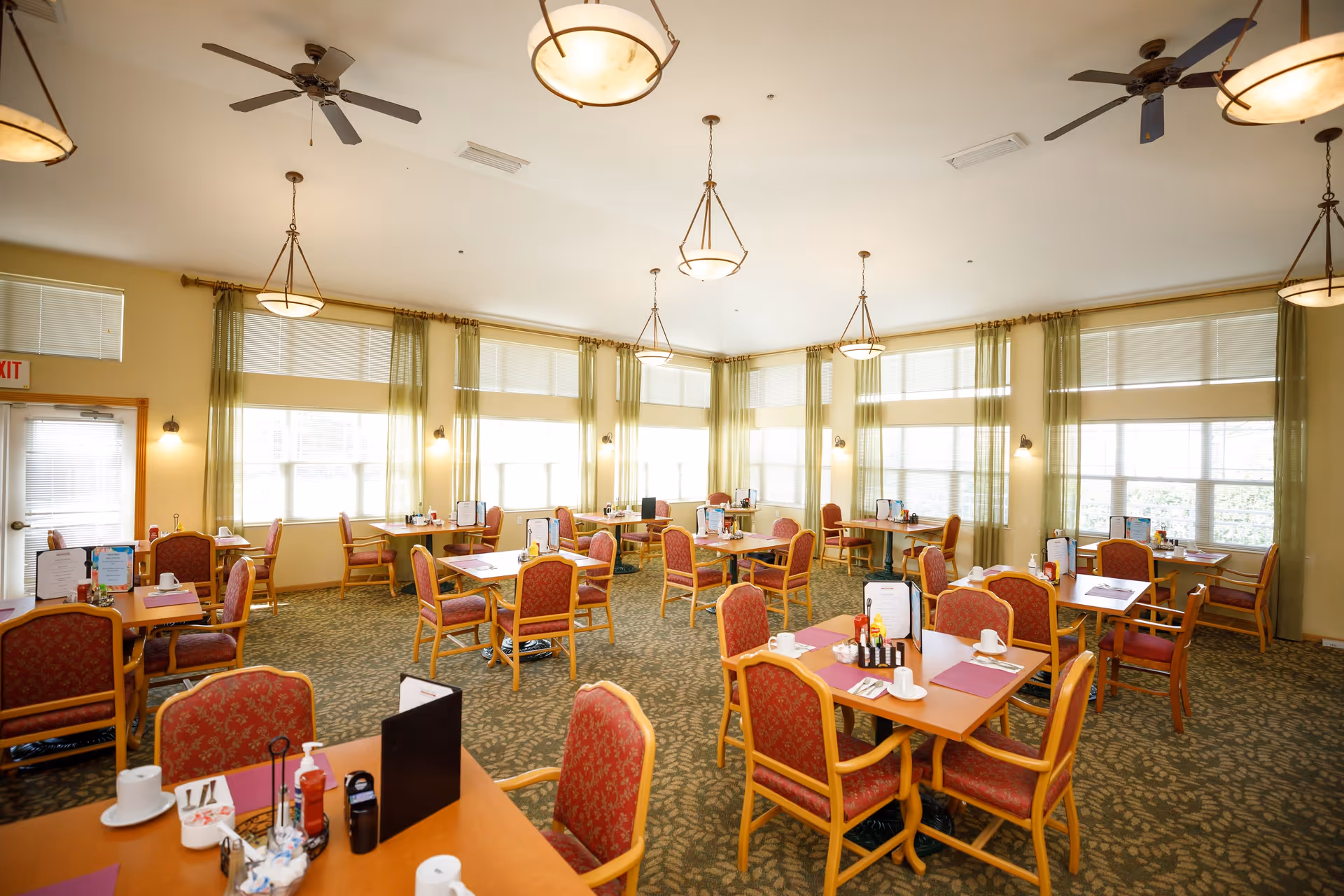 A bright dining room in a senior living facility with multiple wooden tables and red cushioned chairs arranged neatly. The room has large windows with green curtains allowing natural light to fill the space. Ceiling fans and hanging light fixtures are visible on the ceiling. Tables are set with menus, cups, and condiments.