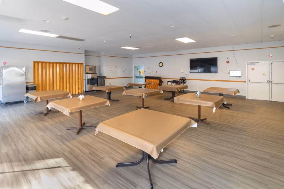 Spacious dining/activity room with several tables covered in brown tablecloths, a wall-mounted TV, and wood-look flooring.
