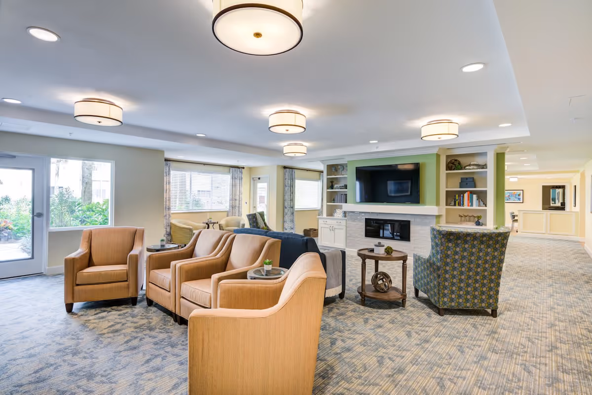 A spacious and well-lit living room area in a senior living facility with multiple comfortable armchairs arranged around small tables. The room features a large flat-screen TV mounted above a modern fireplace, built-in shelves with decorative items and books, large windows with curtains, and soft overhead lighting fixtures. The carpet has a subtle pattern, and there is a door leading outside on the left side.