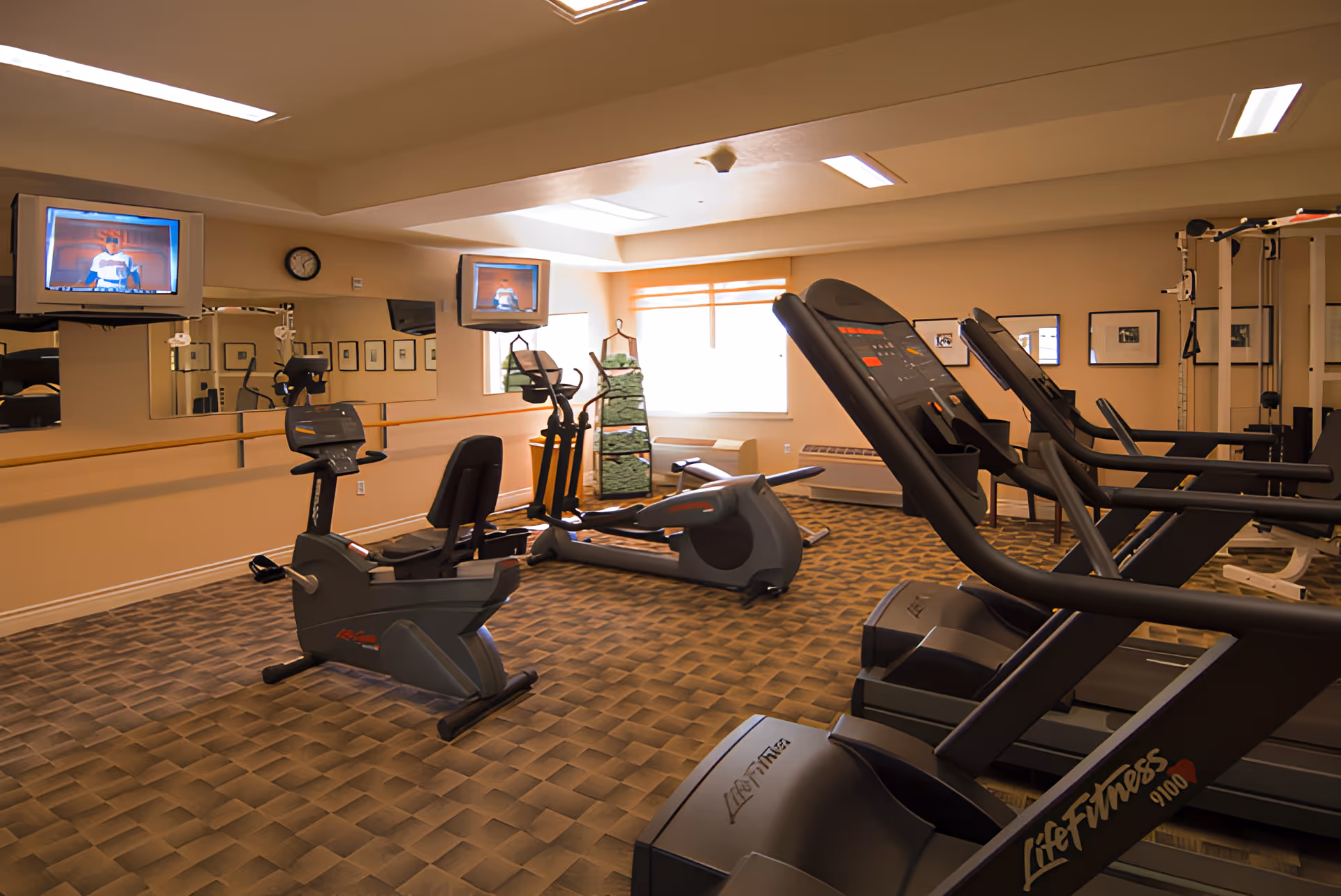 A fitness room with exercise equipment including treadmills, stationary bikes, and elliptical machines. The room has a patterned carpet, beige walls, a large mirror on one wall, two televisions mounted above the mirror, and a window letting in natural light.