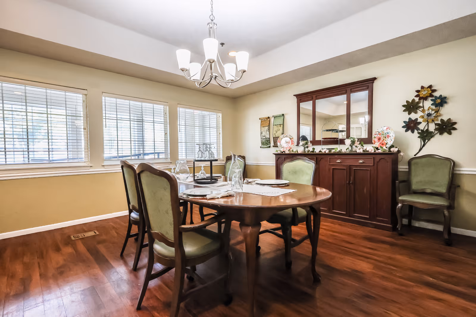 A dining room with a wooden oval table set with plates, glasses, and napkins. Four green upholstered chairs surround the table. There is a wooden sideboard with a large mirror above it, decorated with plates and floral arrangements. Three windows with blinds allow natural light into the room, and a chandelier with five lights hangs from the ceiling. The floor is wooden and the walls are painted in a light beige color with a white chair rail.