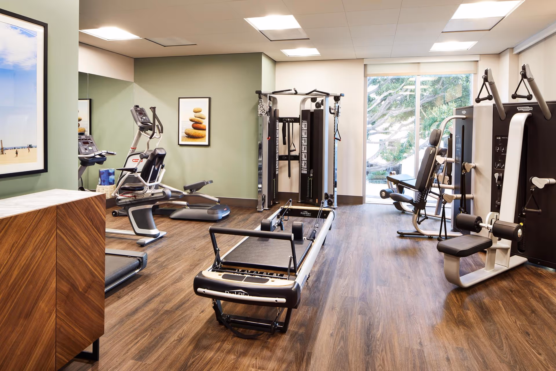 A well-lit fitness room with various exercise equipment including a rowing machine, elliptical trainer, and strength training machines. The room has wood flooring, light green walls, framed artwork, and a large window showing greenery outside.