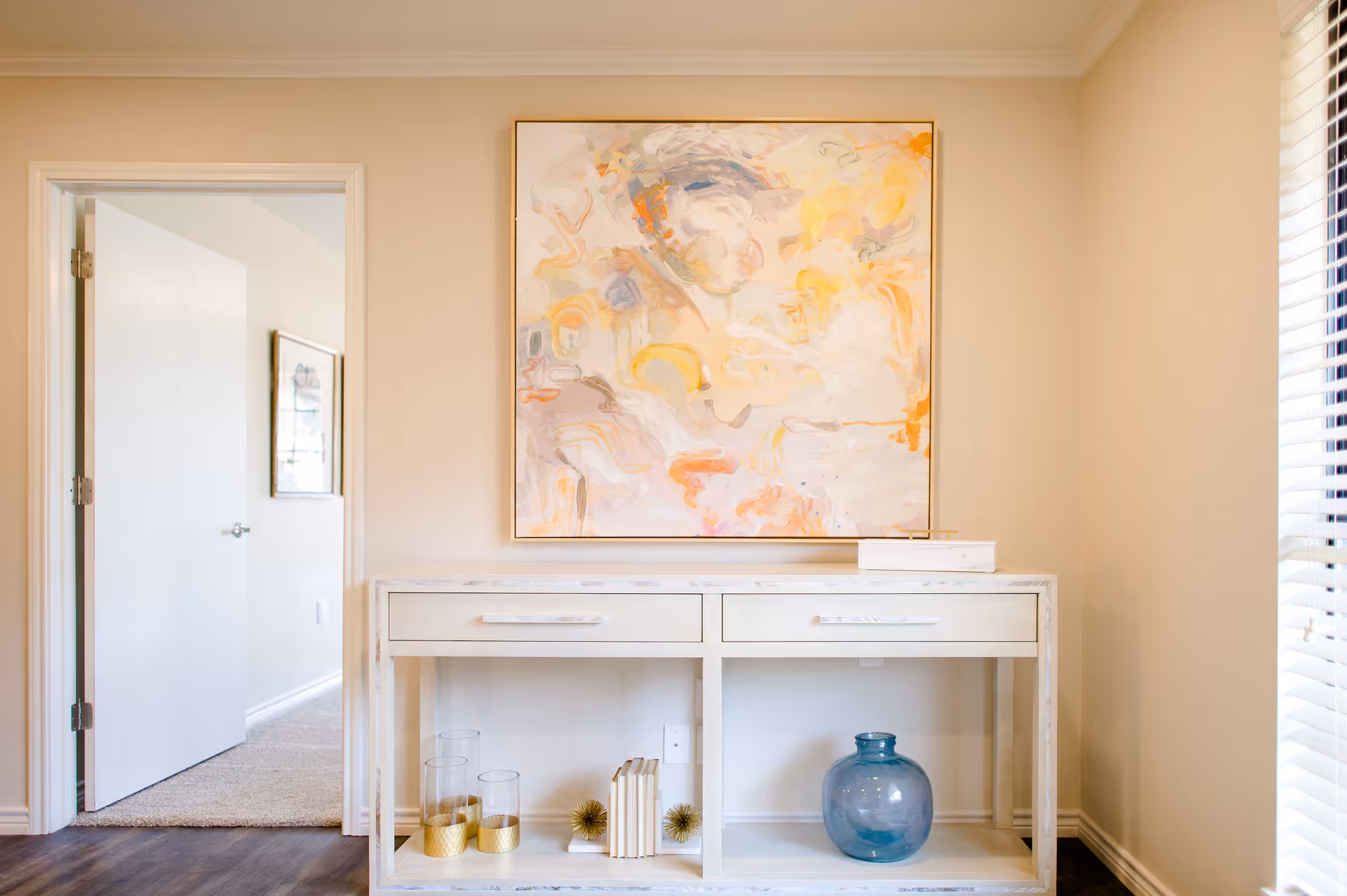 A light-colored console table with decorative vases and books beneath an abstract painting in a bright interior room.