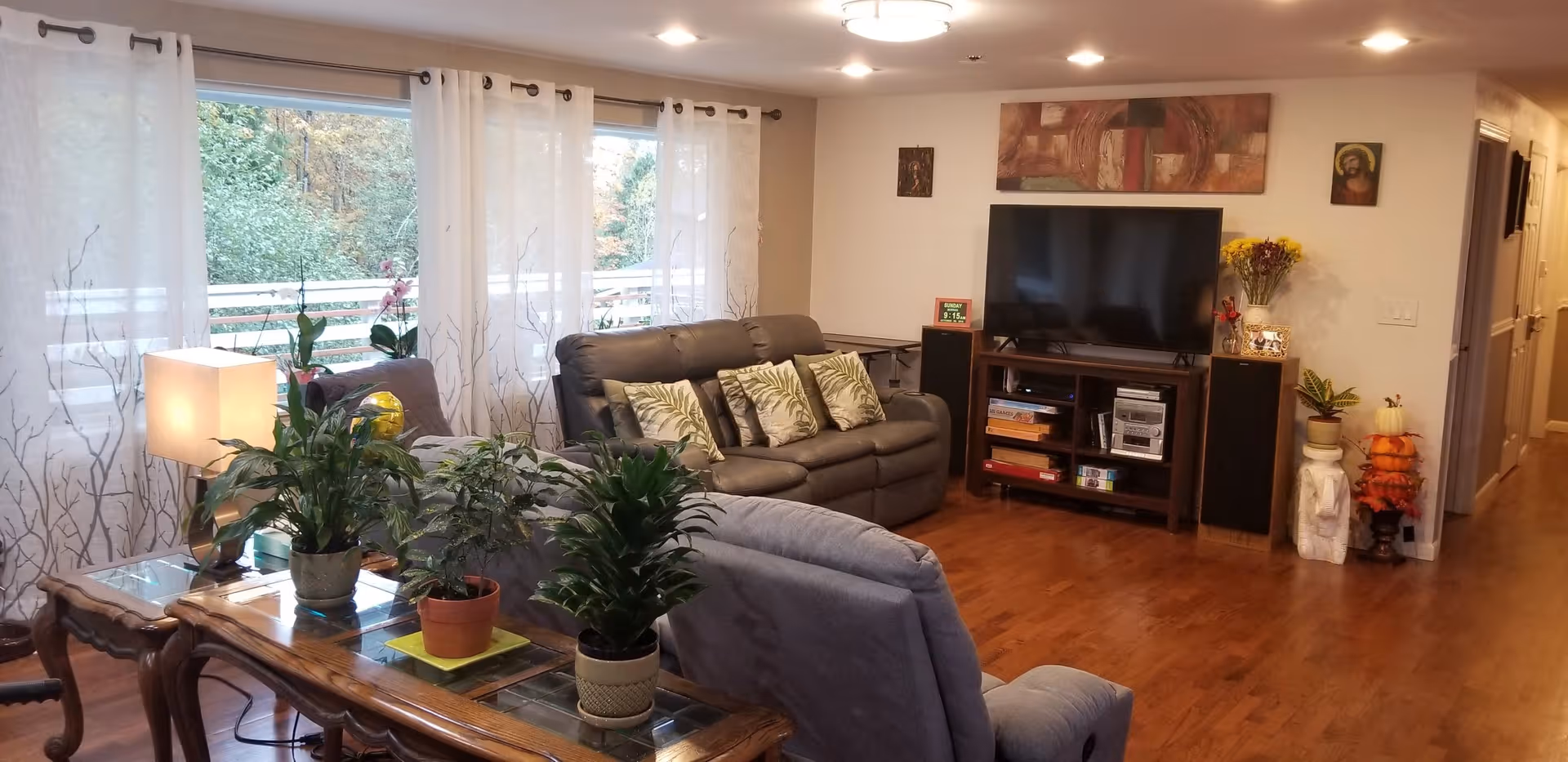 A cozy living room with wooden flooring, a large window with sheer white curtains, a gray recliner chair, a dark gray sofa with patterned cushions, a wooden coffee table with several potted plants, a TV on a wooden stand with speakers on each side, and decorative artwork on the walls.