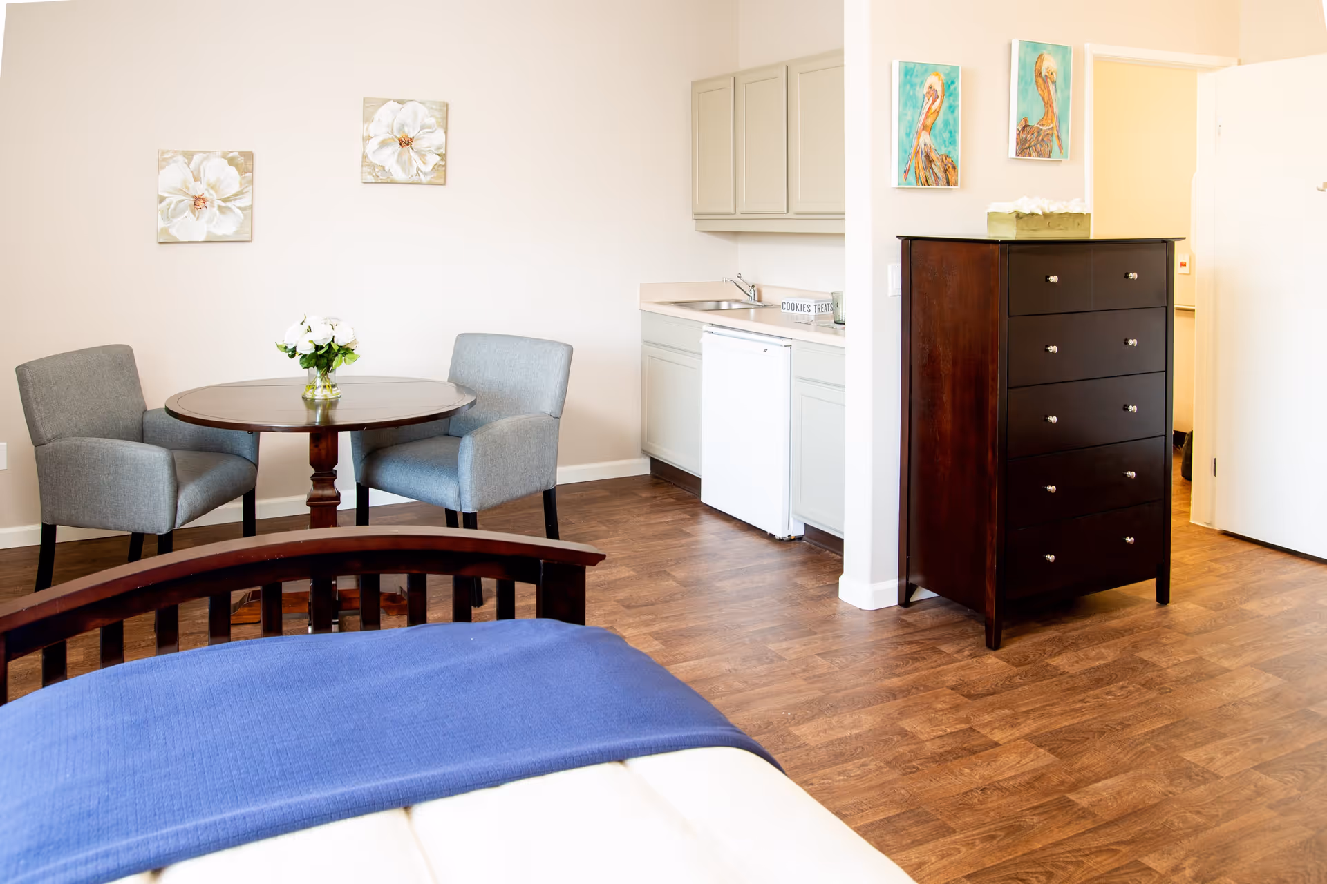 A bright senior living room featuring a wooden bed with a blue blanket, a round wooden table with two gray upholstered chairs, a small kitchenette with a sink and mini refrigerator, and a dark wooden chest of drawers. The walls are decorated with floral and bird paintings, and the floor is wood.