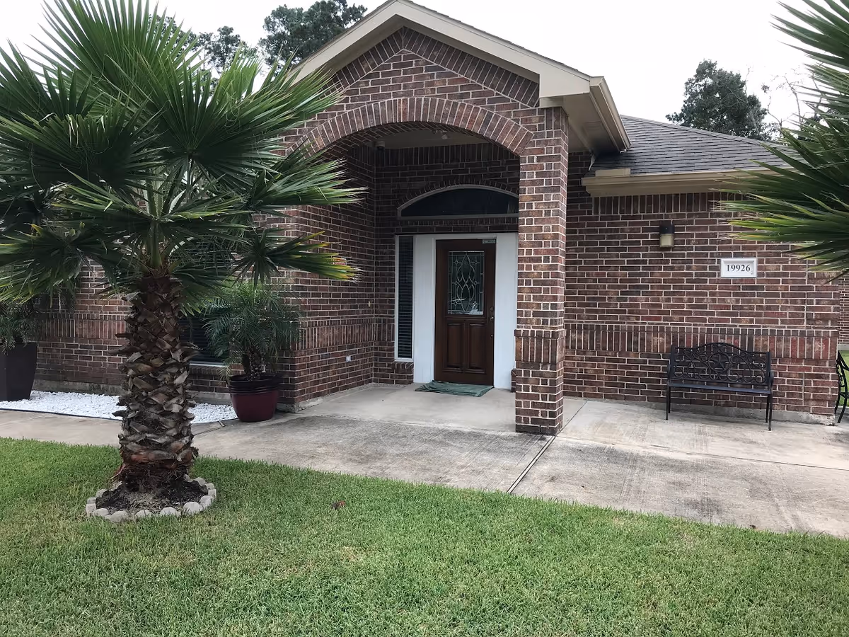 Front exterior view of a brick building with a covered entrance, a wooden door with decorative glass, two potted plants, a small palm tree in the lawn, a black metal bench, and a house number plaque reading 19926.