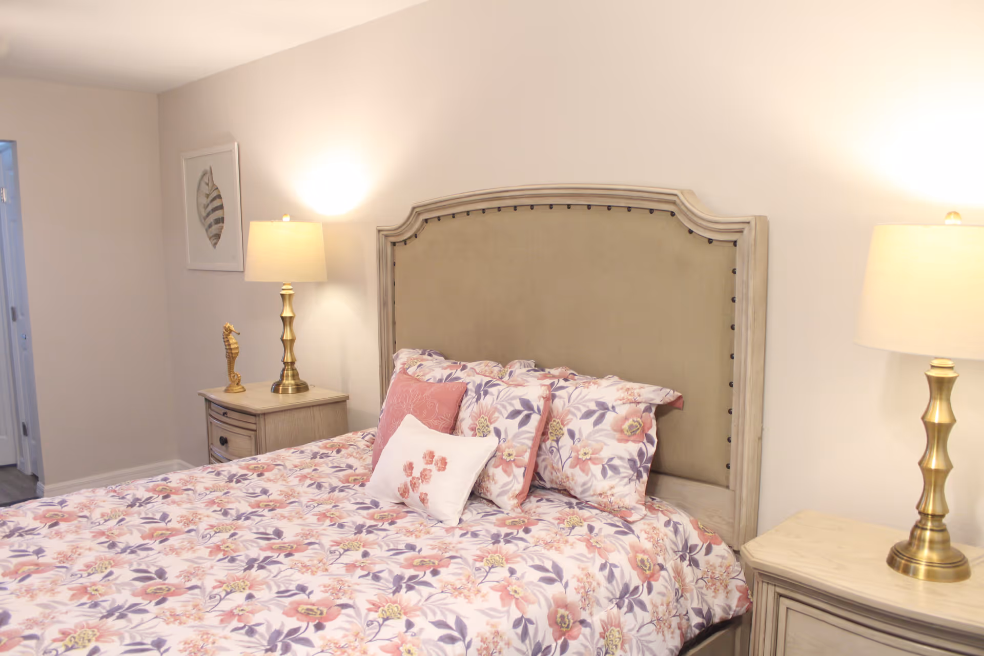 Light, airy bedroom with a floral comforter on a bed with an upholstered headboard, flanked by two nightstands and brass lamps.