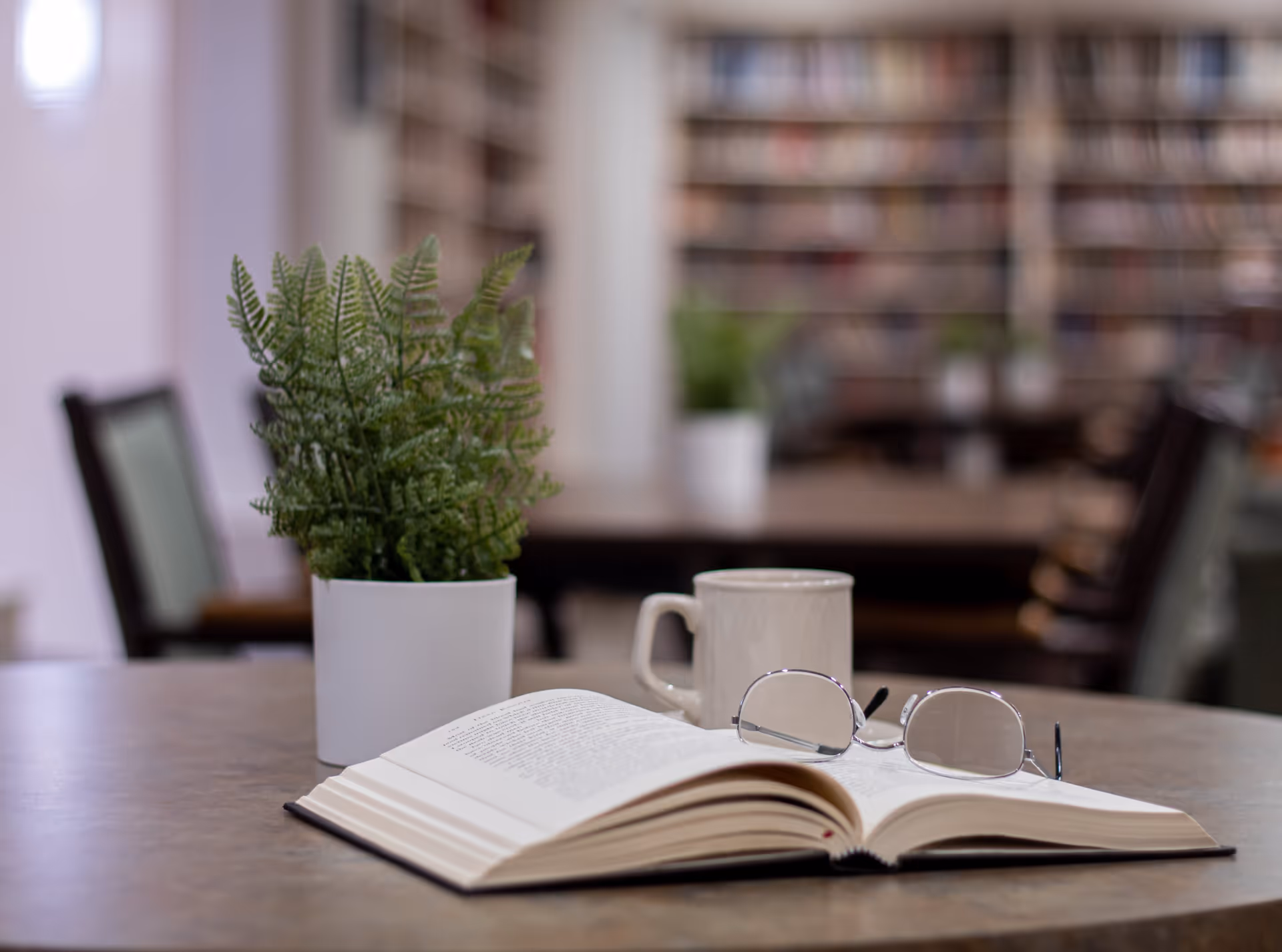 A close-up view of a round table with an open book, a pair of reading glasses resting on the book, a white coffee mug, and a small potted fern plant. In the background, there are blurred bookshelves and chairs, suggesting a library or reading area.