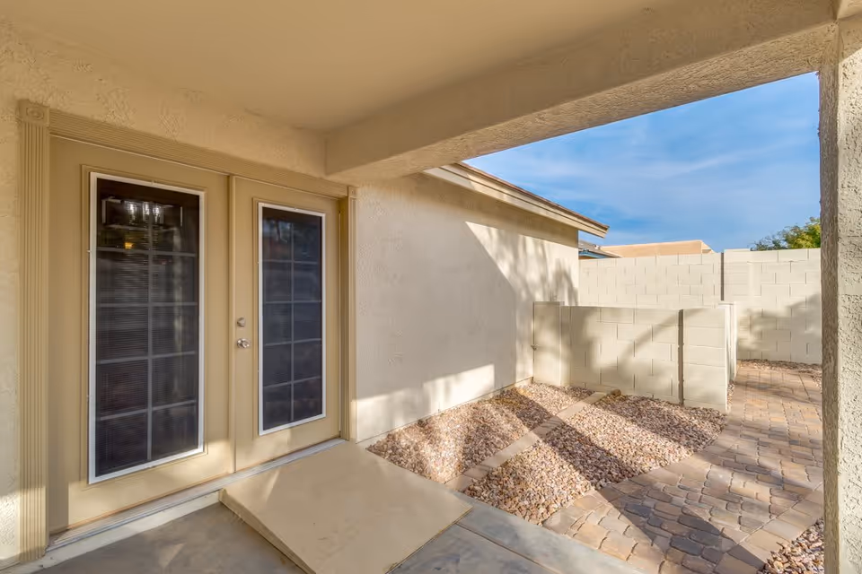 Covered patio area with beige double French doors leading inside. The patio floor is concrete with a small ramp, and the surrounding area features a stone pathway and gravel landscaping enclosed by a light-colored block wall under a clear blue sky.