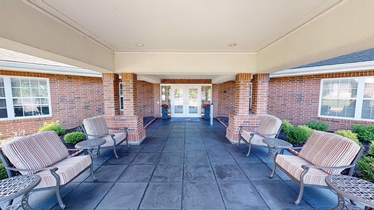 Covered outdoor seating area with four cushioned chairs and three small round tables, brick walls, windows with white blinds, and double glass doors at the far end.