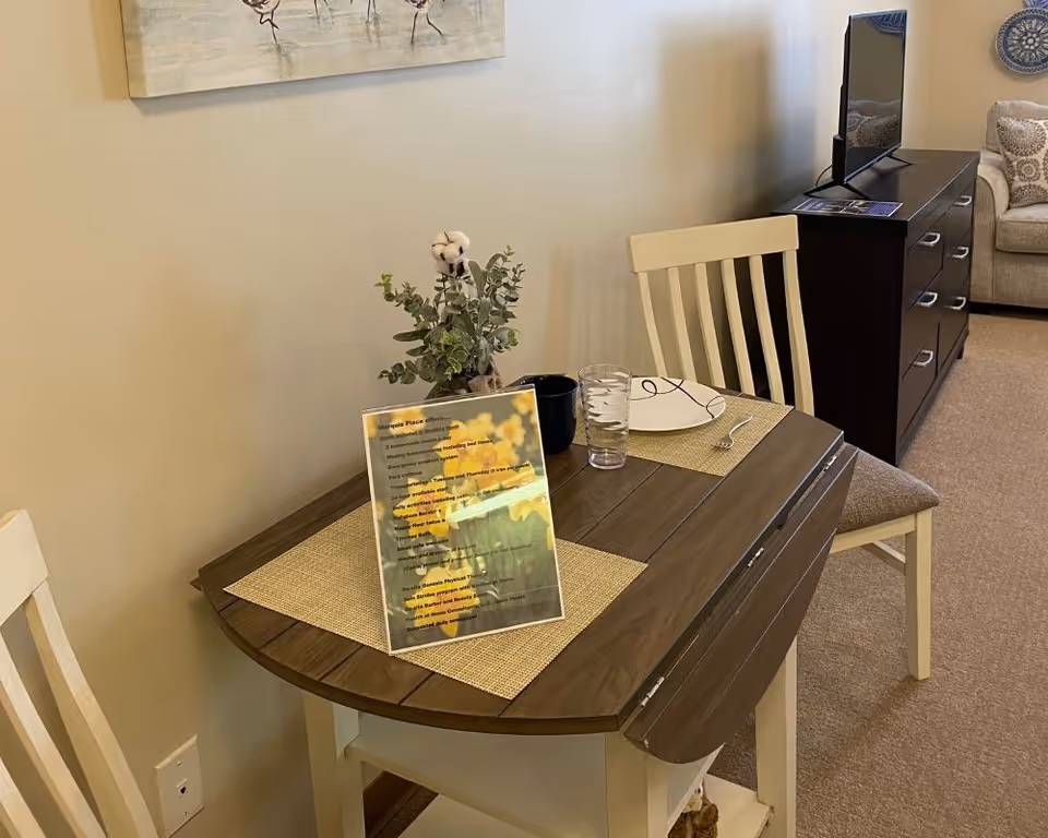 Small dining table set for two with placemats, a framed information sign, and chairs in a living area with a TV and dresser in the background.