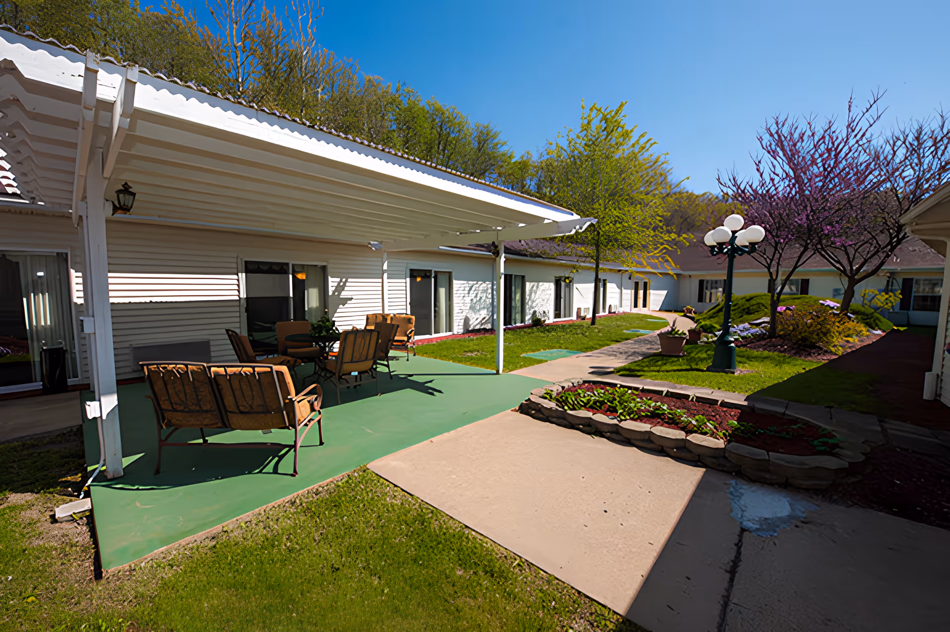 Outdoor patio area at a senior living facility with green flooring, several cushioned chairs and tables under a white pergola. The patio is surrounded by a garden with trees, flowering plants, and a lamppost with multiple globe lights. The building has white siding and multiple windows and doors facing the patio.