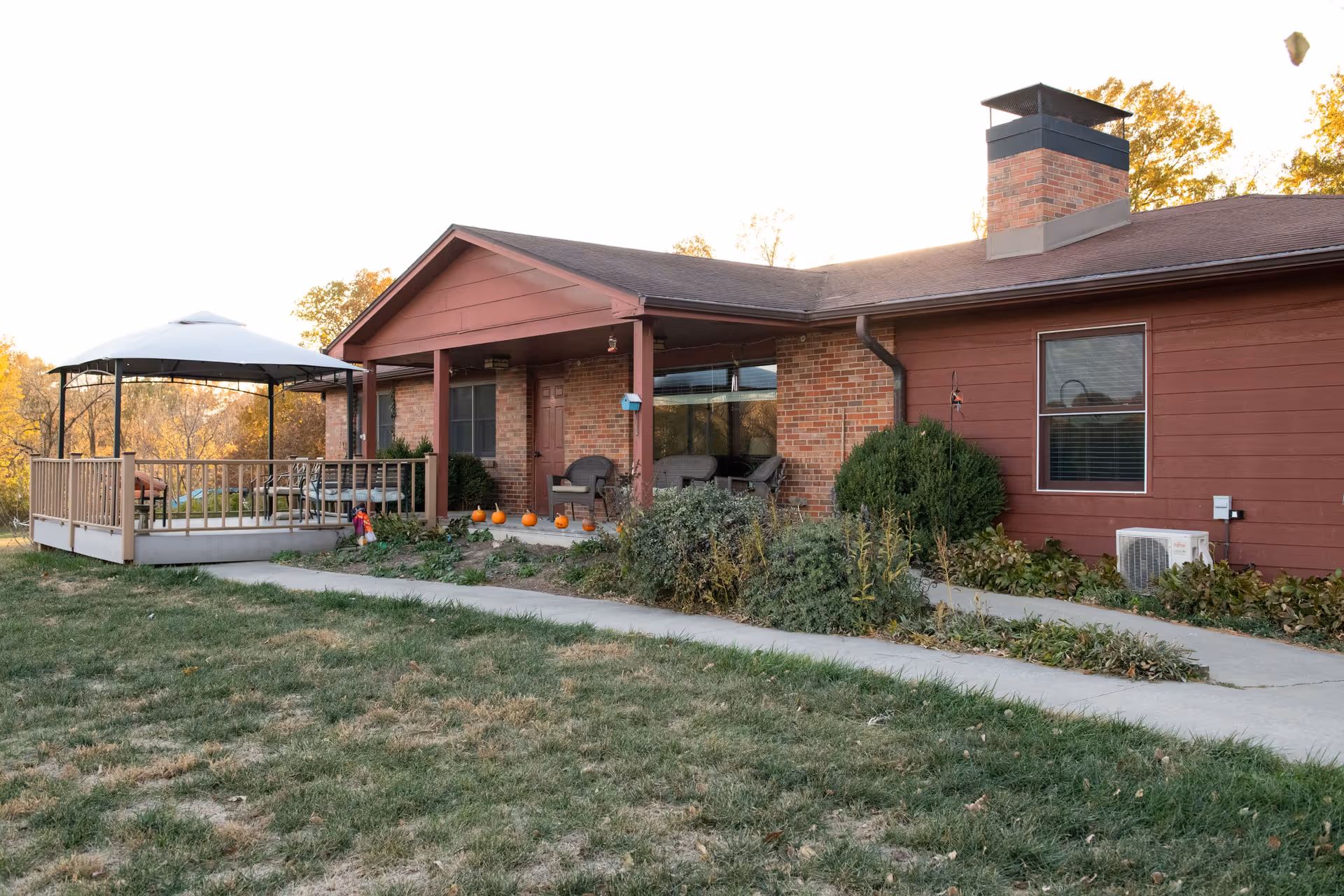 Exterior view of a single-story senior living facility building with red siding and brick accents. The building features a covered porch with chairs and a small garden area with pumpkins placed along the edge. There is a gazebo with a white canopy on a raised wooden deck to the left, and a concrete walkway leads up to the porch. Trees with autumn foliage are visible in the background.