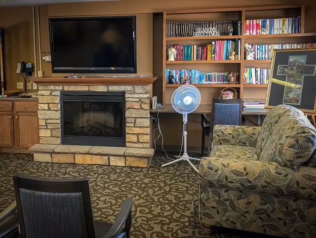 A cozy living room area featuring a stone fireplace with a mounted flat-screen TV above it. To the right of the fireplace is a wooden bookshelf filled with books and decorative items, including a 'Season's Greetings' sign. A standing fan is positioned in front of the bookshelf. There is a patterned armchair on the right side and a dark wooden chair partially visible in the foreground. The room has a patterned carpet and wooden cabinetry on the left side.
