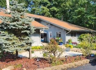 Exterior view of a single-story building with gray siding and a reddish-brown roof, surrounded by landscaped garden beds with various plants and shrubs, and a large evergreen tree on the left side.