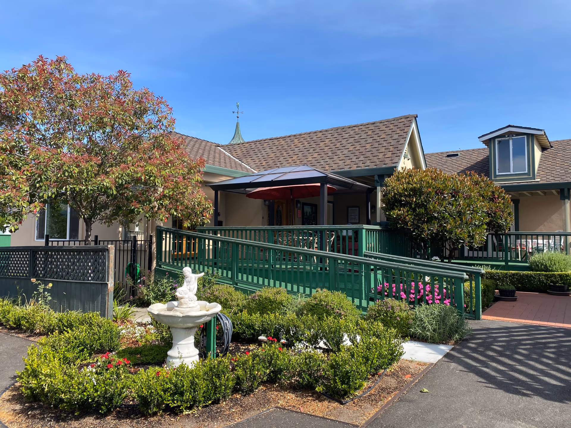 Exterior view of Bella Vista Village II showing a single-story building with a brown shingled roof, green railings, and a ramp leading to the entrance. There are well-maintained bushes, a small white fountain with a statue, and flowering plants in the garden area under a clear blue sky.