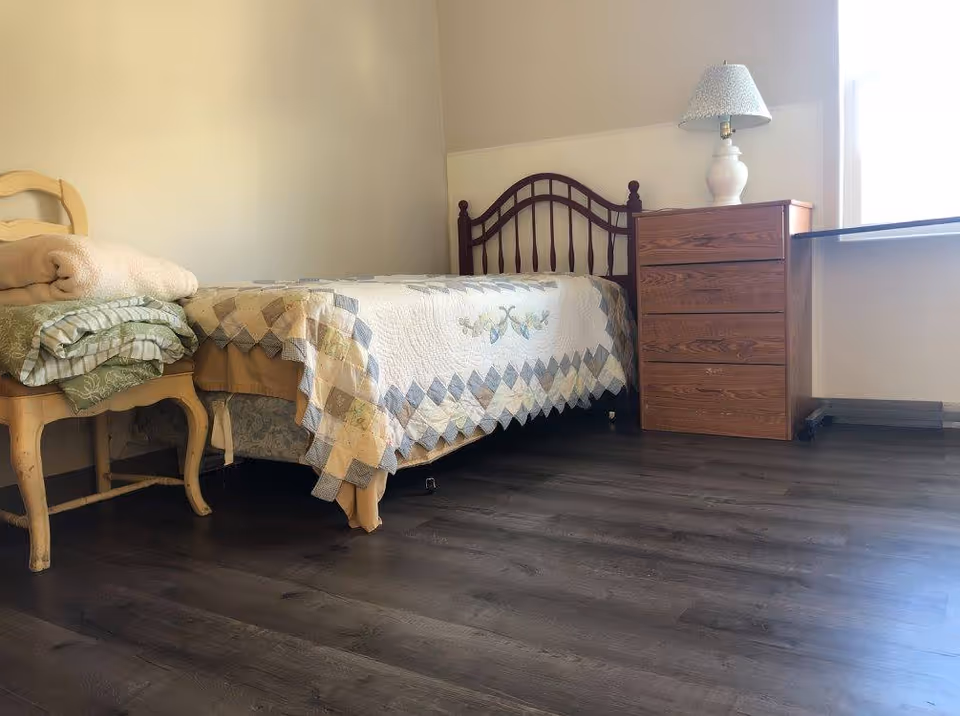 Small bedroom with a single bed covered by a patchwork quilt, wooden headboard, dresser with a lamp, a chair holding folded blankets, and dark laminate flooring.