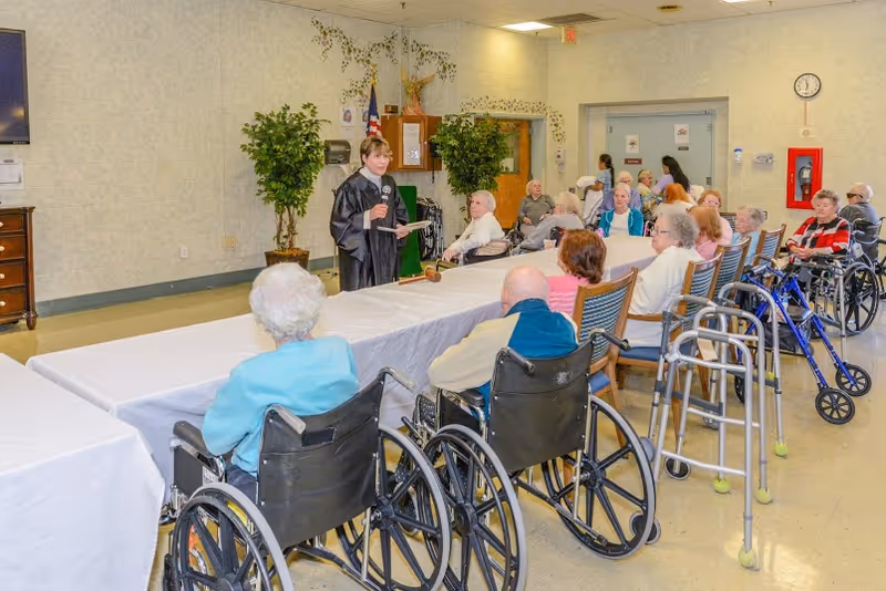 A group of elderly people, many in wheelchairs, seated around long tables covered with white tablecloths in a large room. A woman in a black robe stands at the front holding a microphone and papers, addressing the group. The room has light-colored walls, plants, a clock, and a fire extinguisher on the wall.