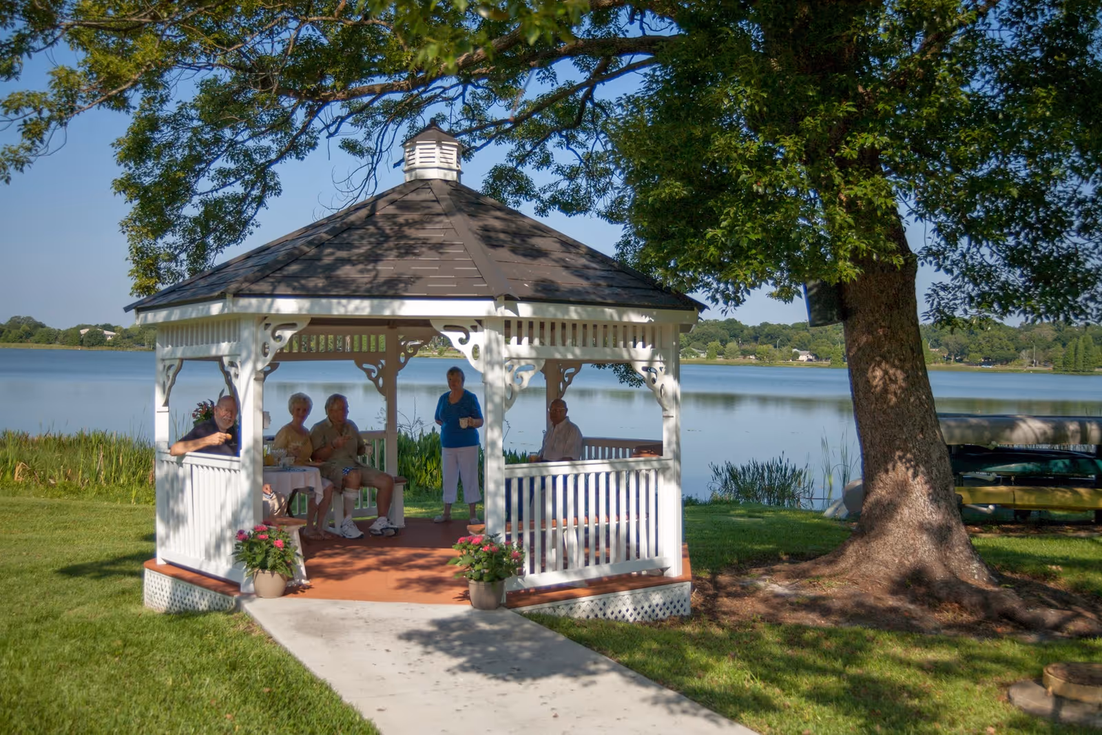 A white lakeside gazebo with several seniors sitting and standing inside under a large tree.
