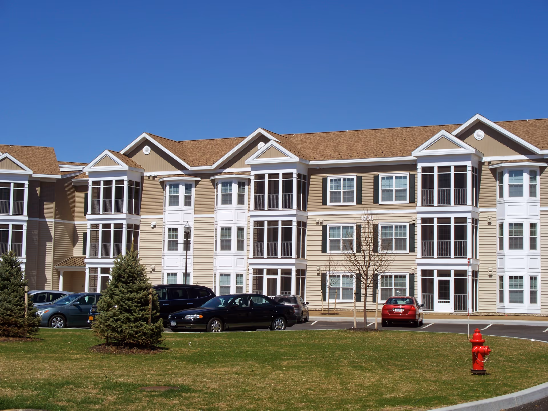 Exterior view of a multi-story residential building with beige siding, white trim, and multiple large windows. Several cars are parked in front of the building on a paved parking lot. There are small evergreen trees and a red fire hydrant on a grassy area in the foreground under a clear blue sky.