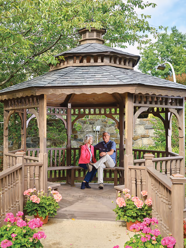 An elderly couple sitting and smiling together inside a wooden gazebo surrounded by greenery and pink flowers in a garden setting.