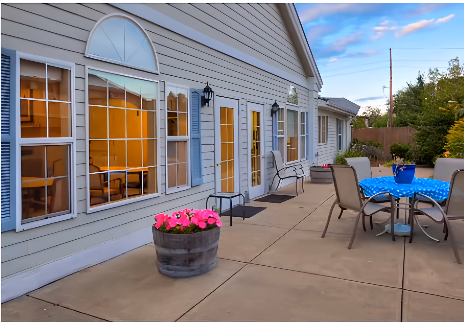 Outdoor patio area of a senior living facility with a concrete floor, a round table covered with a blue tablecloth, surrounded by four chairs. There are potted plants with pink flowers and green shrubs along the side of the building. The building has light-colored siding, multiple windows with blue shutters, and glass doors. The sky is partly cloudy with a hint of sunset.