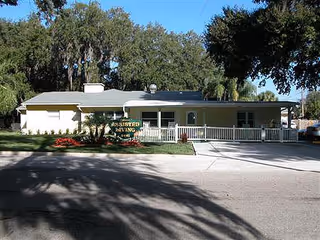 Single-story building with a light-colored exterior surrounded by trees and landscaping. A sign in front reads 'Assisted Living'. The building has a covered entrance and a paved driveway.