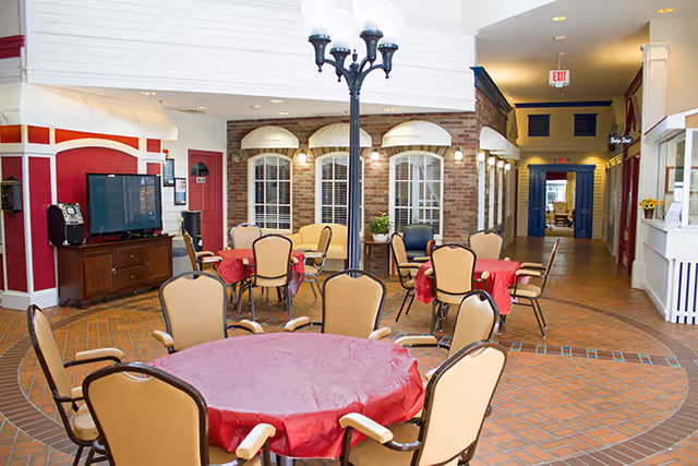 Interior view of a common area in Buckingham Heights Memory Care featuring round tables covered with red tablecloths and surrounded by beige chairs. The space has a brick floor and walls designed to resemble a small town street with storefront windows and doors. A black streetlamp-style light fixture is in the center, and a television is visible on a wooden cabinet to the left.