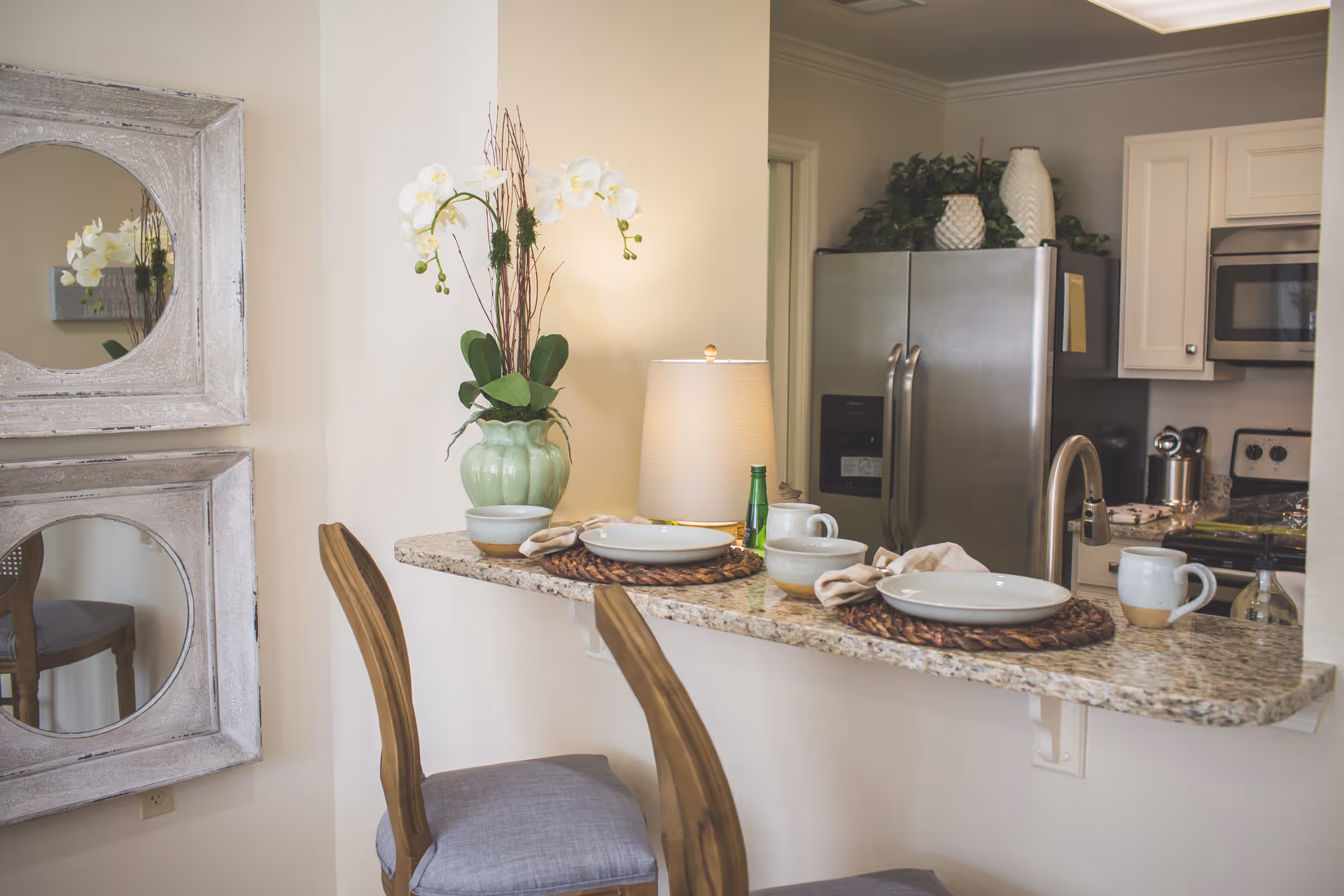 Granite breakfast bar set for two with wooden stools, decorative orchid and lamp, overlooking a kitchen with stainless steel refrigerator and microwave.