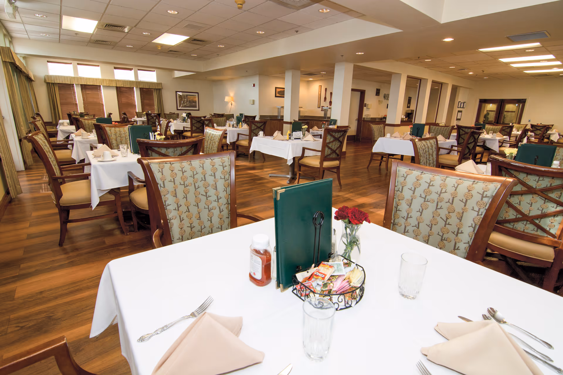 A spacious dining room with multiple tables covered in white tablecloths, each set with beige napkins, glasses, and silverware. The chairs have wooden frames with patterned upholstery. The room has wooden flooring, large windows with blinds, and soft lighting from ceiling fixtures and wall lamps. A table in the foreground has a green menu, a small vase with red flowers, and a condiment basket.