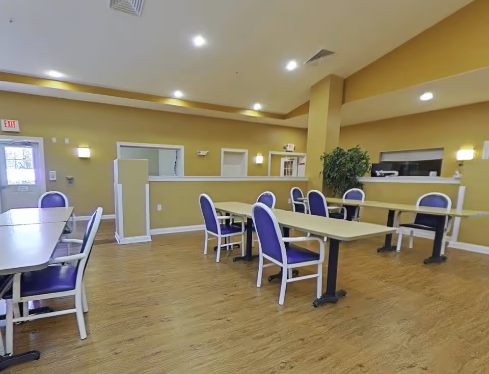 A spacious room with several tables and purple cushioned chairs arranged around them. The walls are painted a warm yellow color, and the floor has a wood-like finish. There are ceiling lights and wall sconces providing illumination. A green plant is placed near the back wall, and there are windows and doors visible in the background.