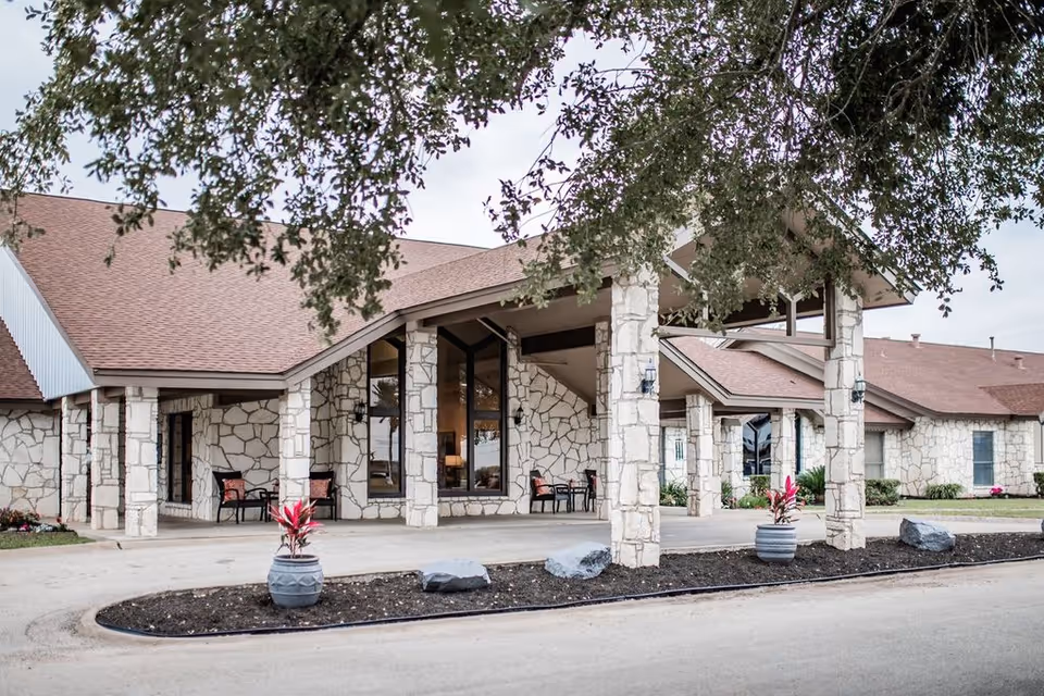 Exterior view of a nursing and rehabilitation center building with stone facade and a covered entrance supported by stone pillars. There are potted plants and outdoor seating visible near the entrance, with a tree branch partially framing the top of the image.