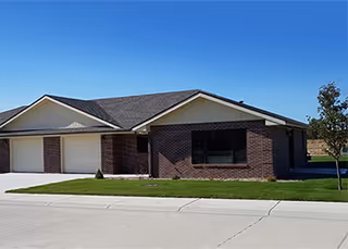 Single-story brick building with a two-car garage, a large window, and a well-maintained lawn under a clear blue sky.