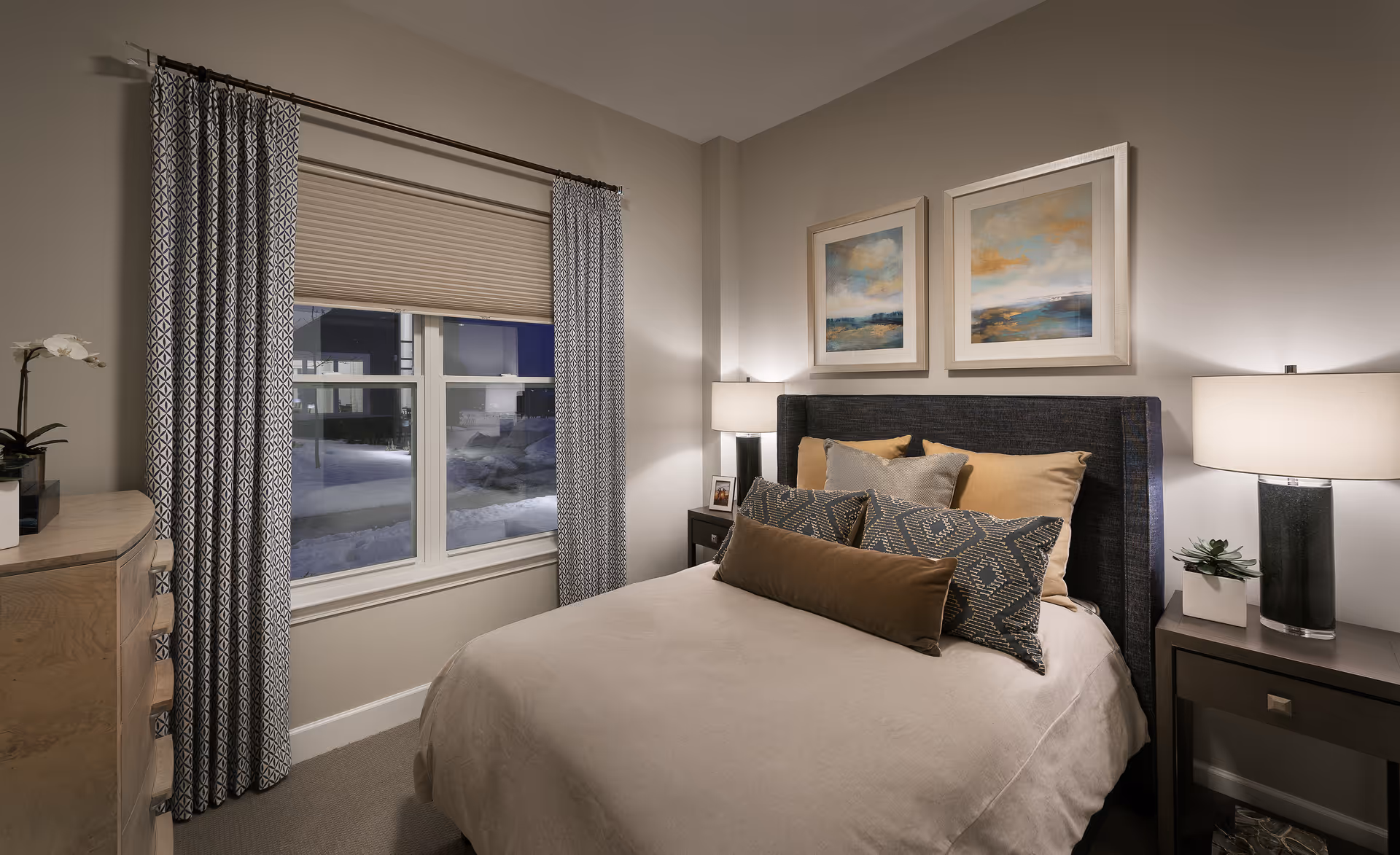 A cozy bedroom with a neatly made bed featuring multiple pillows in neutral tones. The bed has a dark upholstered headboard. On either side of the bed are nightstands with modern lamps and small decorative plants. Above the bed are two framed abstract landscape paintings. A window with patterned curtains and a closed blind is on the left wall, and a wooden dresser with a white orchid plant is partially visible.