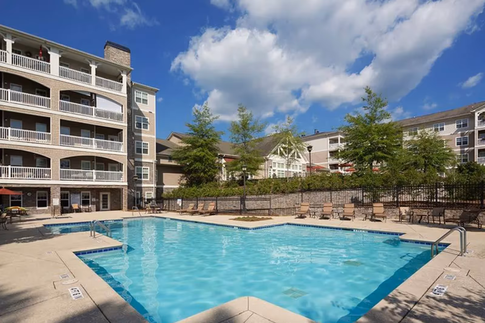 Outdoor swimming pool and patio with lounge chairs in front of a multi-story senior living building under a blue sky.