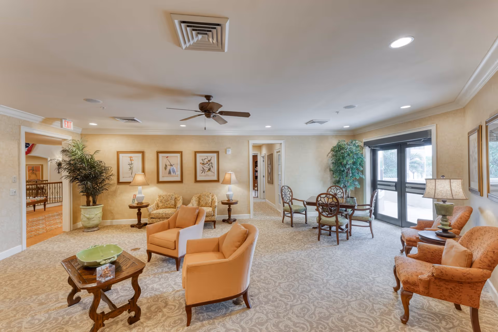 Bright carpeted senior living lounge with upholstered chairs, side tables, framed art, a small table and chairs, and glass doors to the outside.