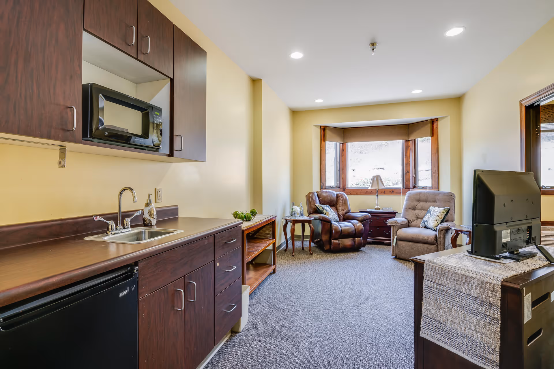 A cozy living area with a kitchenette on the left featuring dark wood cabinets, a microwave, a sink, and a mini refrigerator. The room has beige walls and carpeted floor. At the far end, there are two comfortable armchairs with pillows, a small side table with a lamp, and a large window with wooden trim letting in natural light. A TV is placed on a wooden stand on the right side of the room.