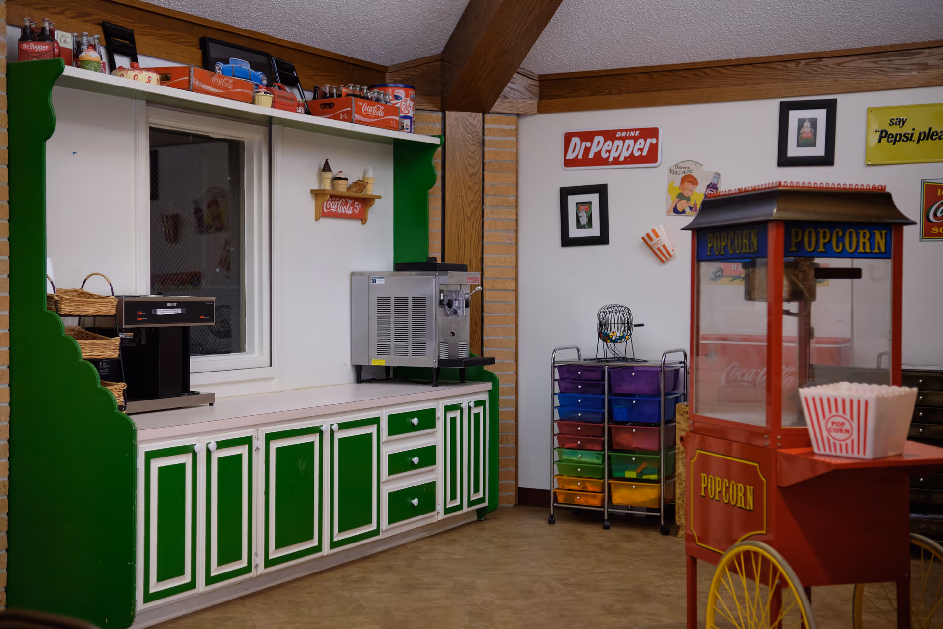 A snack concession area with a red popcorn machine, green paneled cabinets, beverage machines and vintage soda signs on the wall.